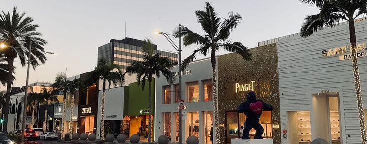 A street scene at dusk showing a row of upscale storefronts, including a prominent Piaget sign, with palm trees, a large black gorilla statue, and a pale evening sky.