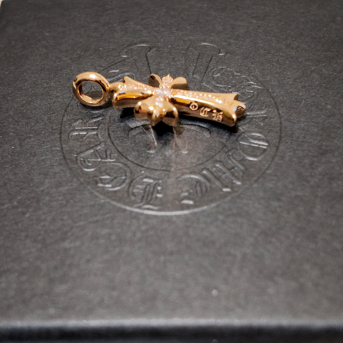 A gold cross pendant with a loop at the top, resting on a dark gray textured surface with an embossed circular logo in the background.