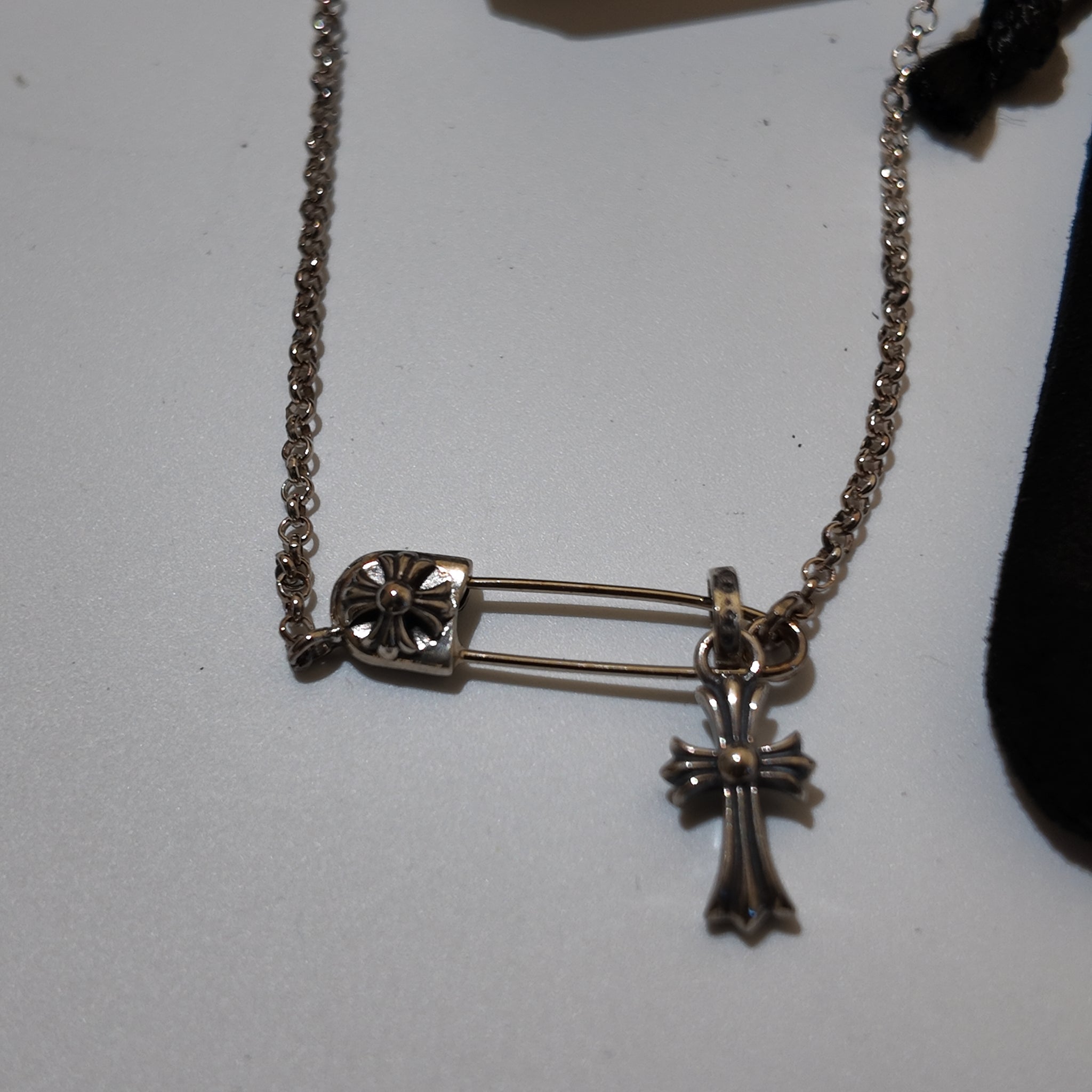 A silver-toned safety pin and cross pendant necklace on a delicate chain, displayed on a light gray surface.