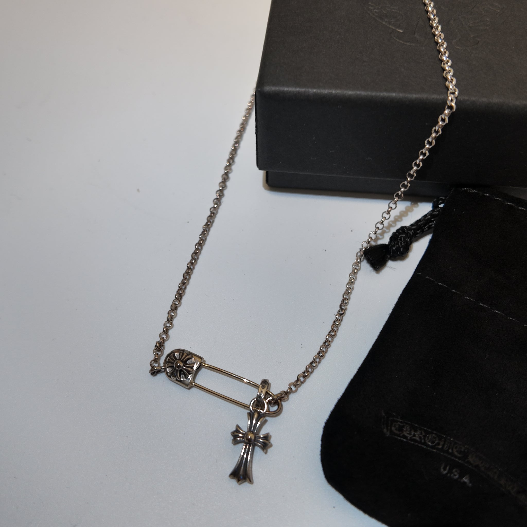 A silver-tone safety pin and cross pendant necklace displayed on a white surface next to a black presentation box and a black velvet pouch with 'Cruel Girls' branding.