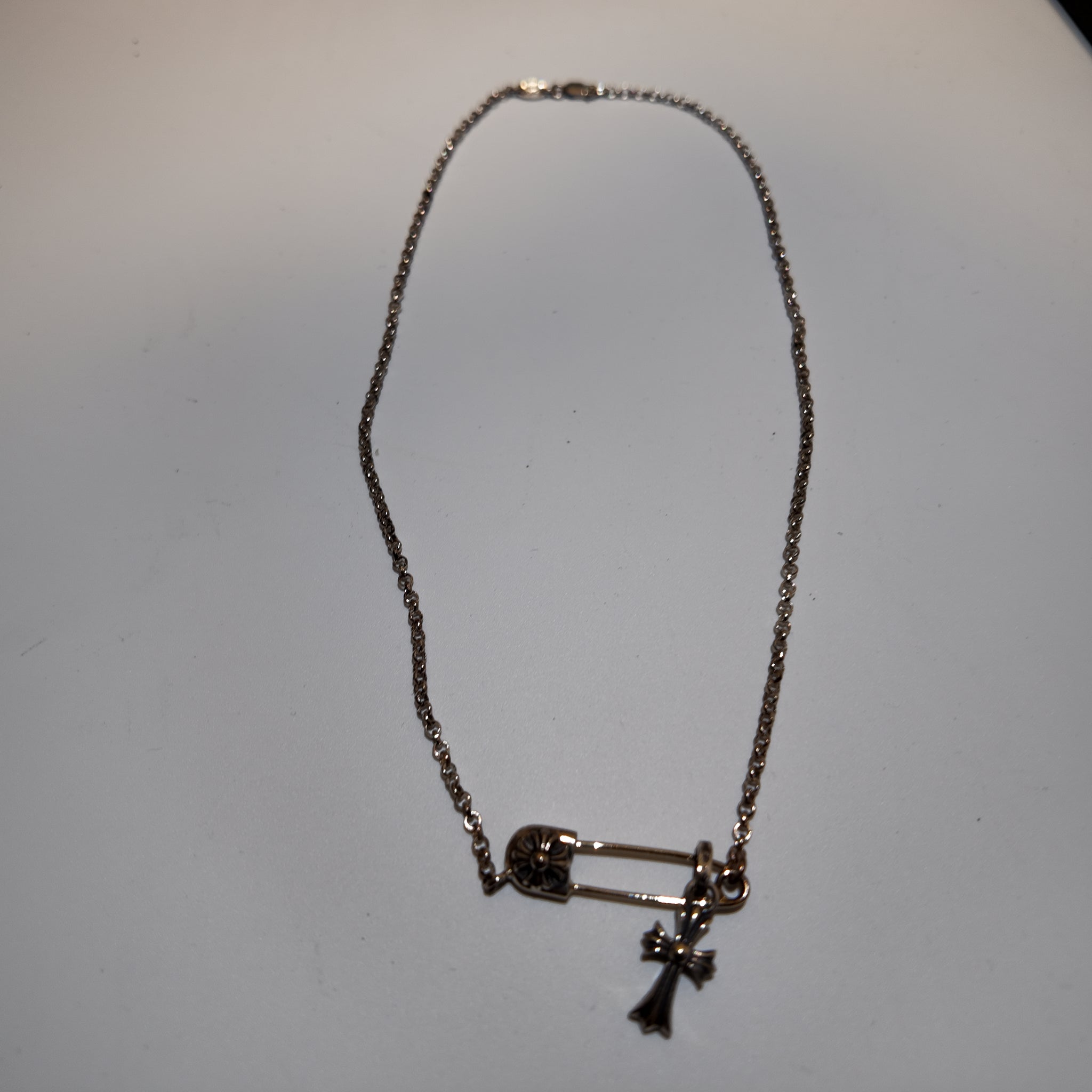 A silver-toned chain necklace featuring a safety pin and cross pendant, displayed on a plain light gray surface.