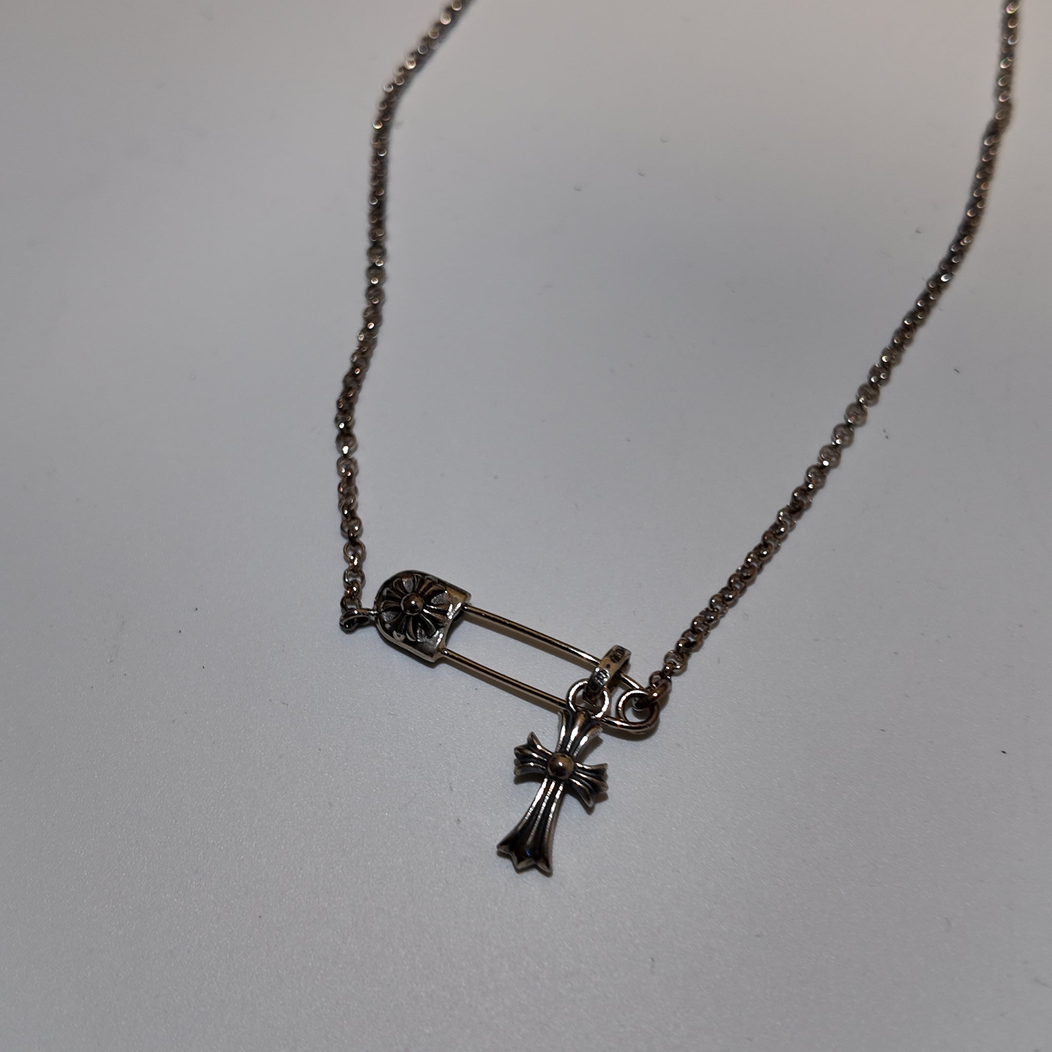 A silver-toned safety pin and cross pendant necklace on a delicate chain, displayed on a plain light gray surface.