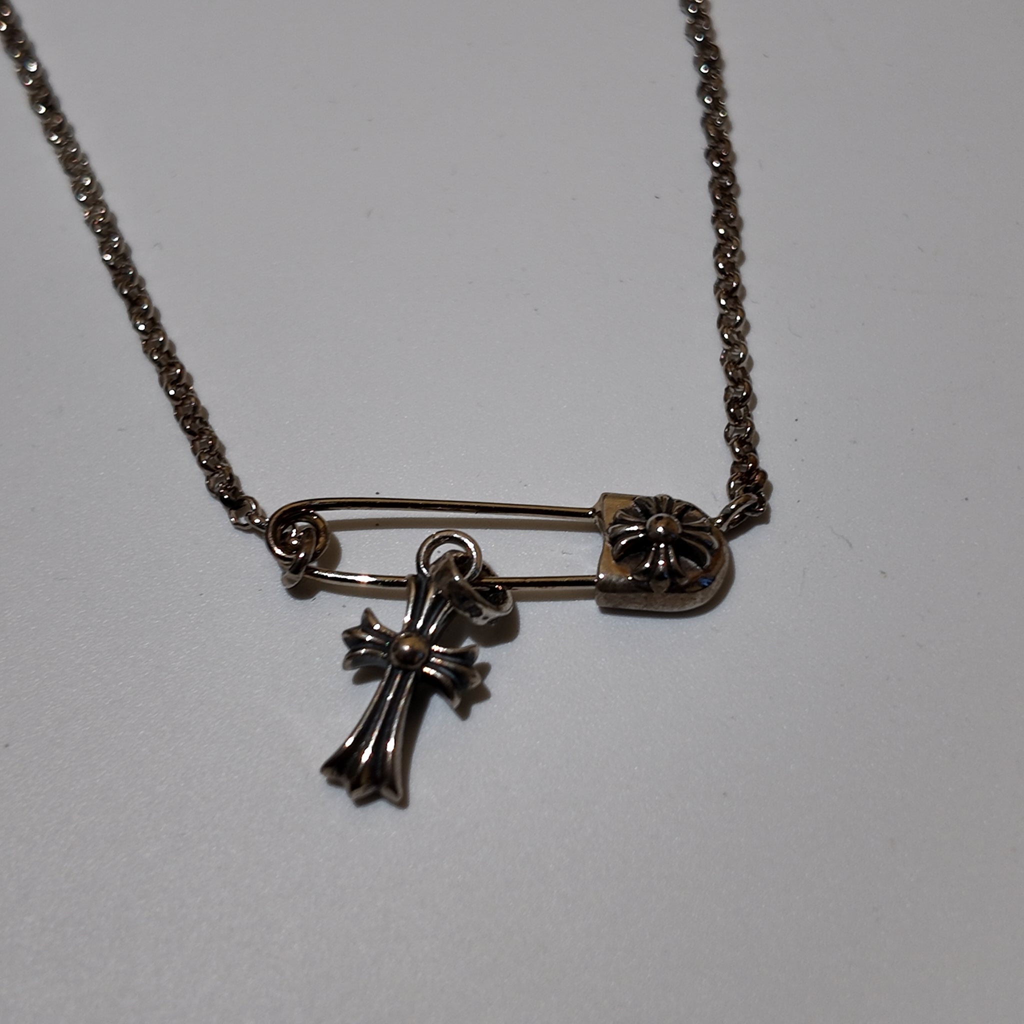 A silver-toned safety pin necklace with a small cross pendant and a flower-shaped clasp, shown on a plain light background.