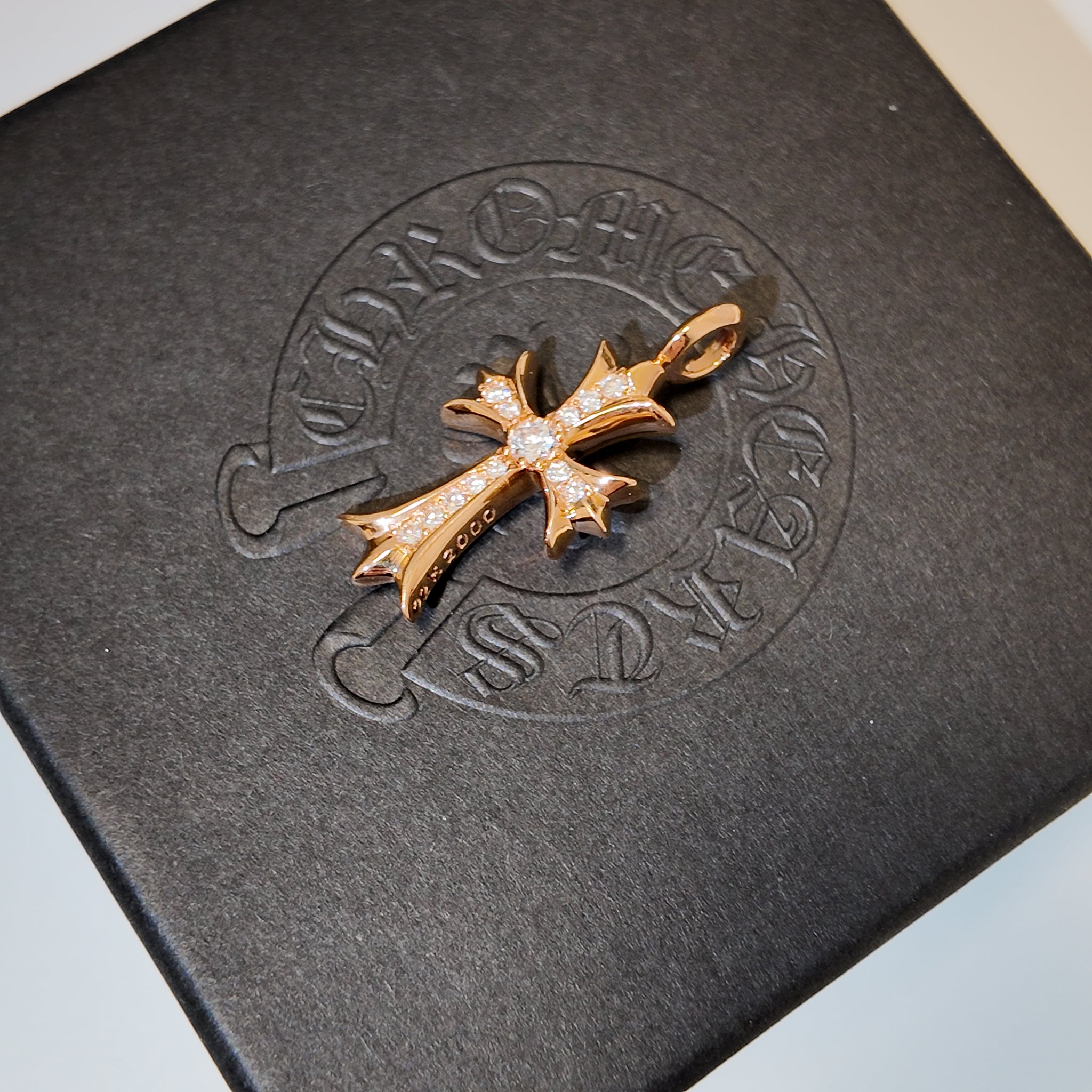 A gold cross pendant paved with small clear stones, displayed on a textured black box embossed with the brand name 'Chap-Lait'.