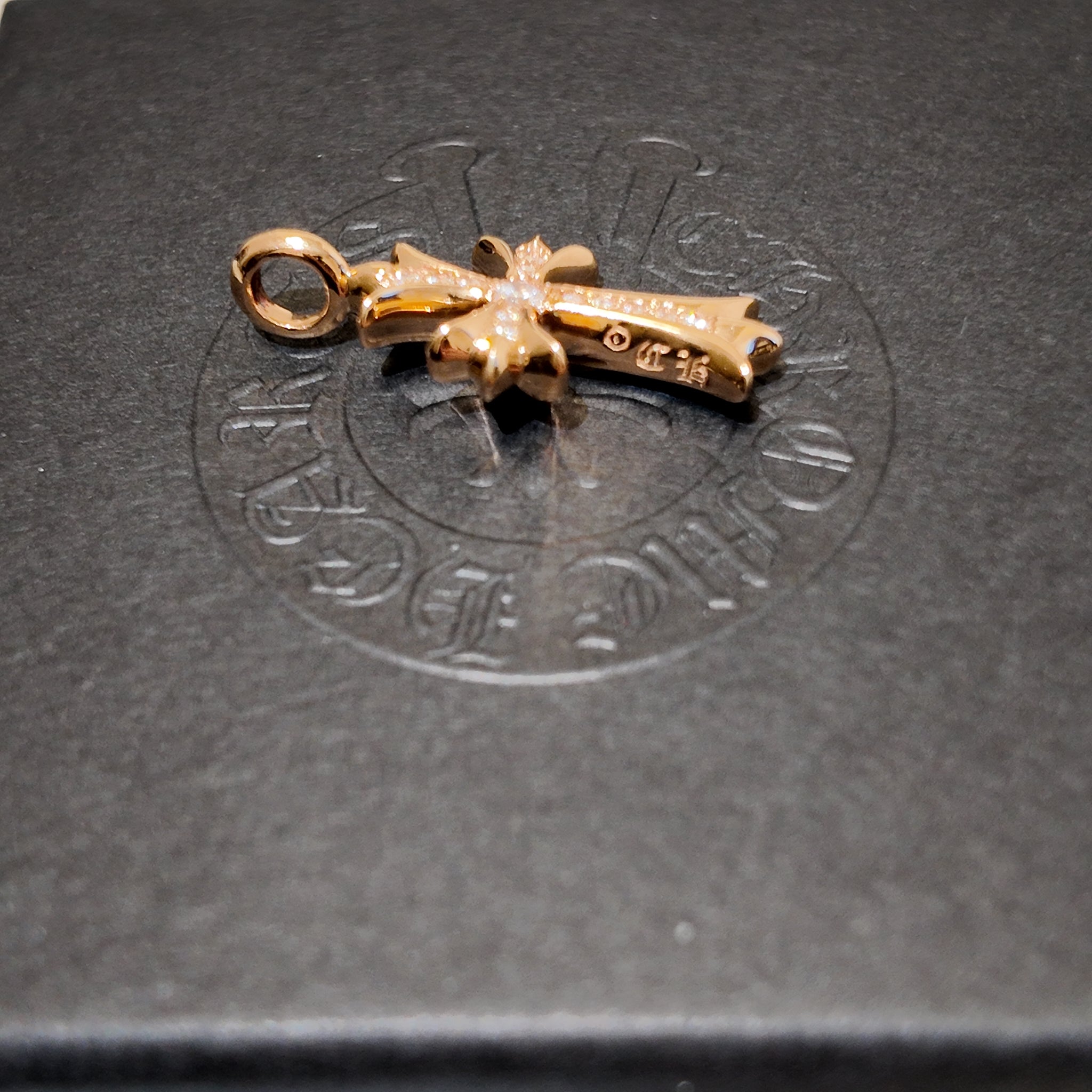 A gold cross pendant with a loop at the top, resting on a dark gray textured surface with an embossed circular logo in the background.