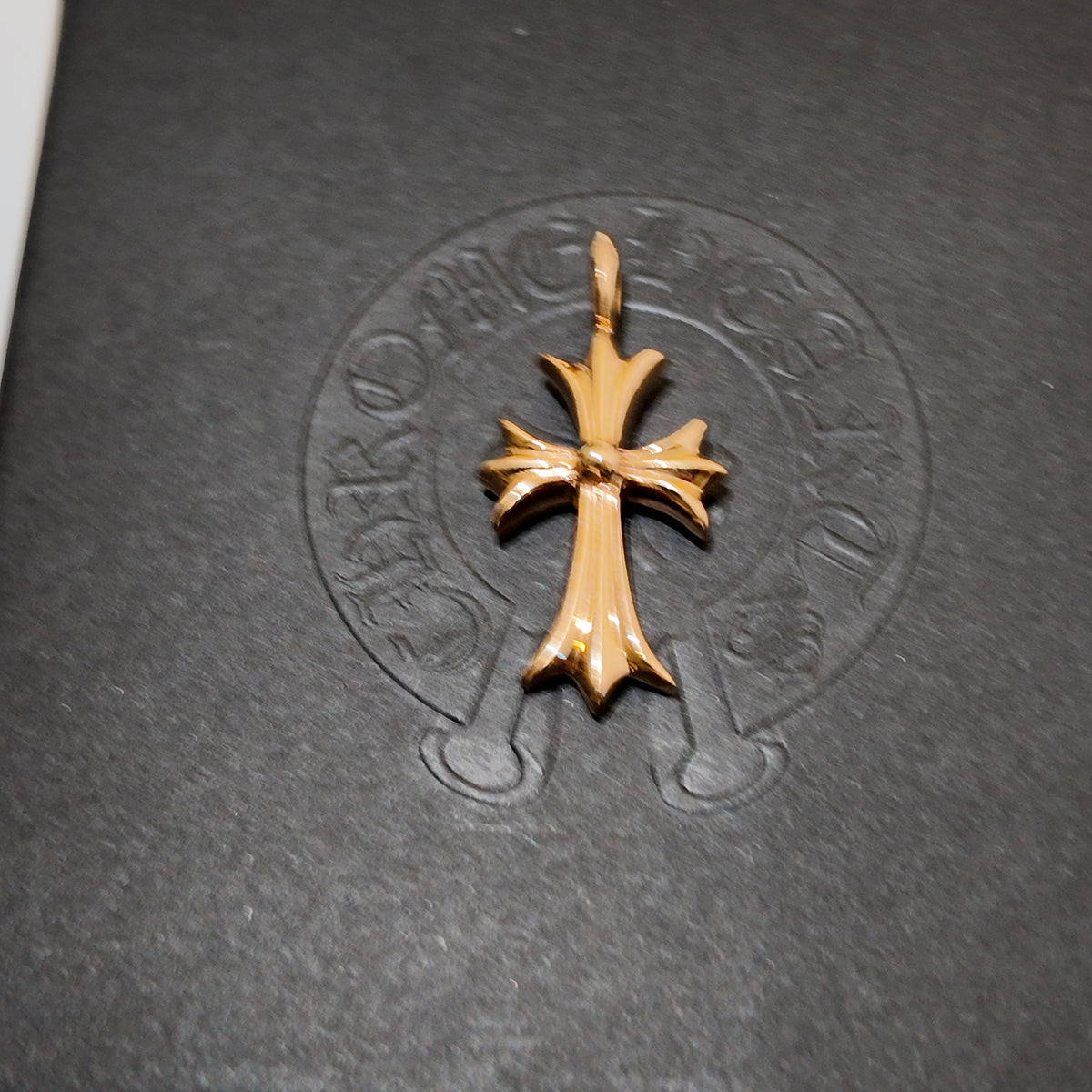 A gold cross pendant with a textured, polished finish, displayed on a dark gray textured surface with an embossed circular logo in the background.