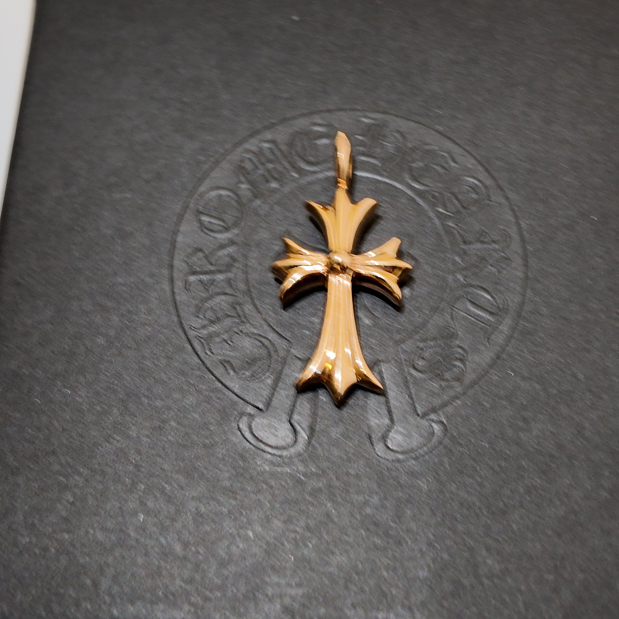 A gold cross pendant with a textured, polished finish, displayed on a dark gray textured surface with an embossed circular logo in the background.