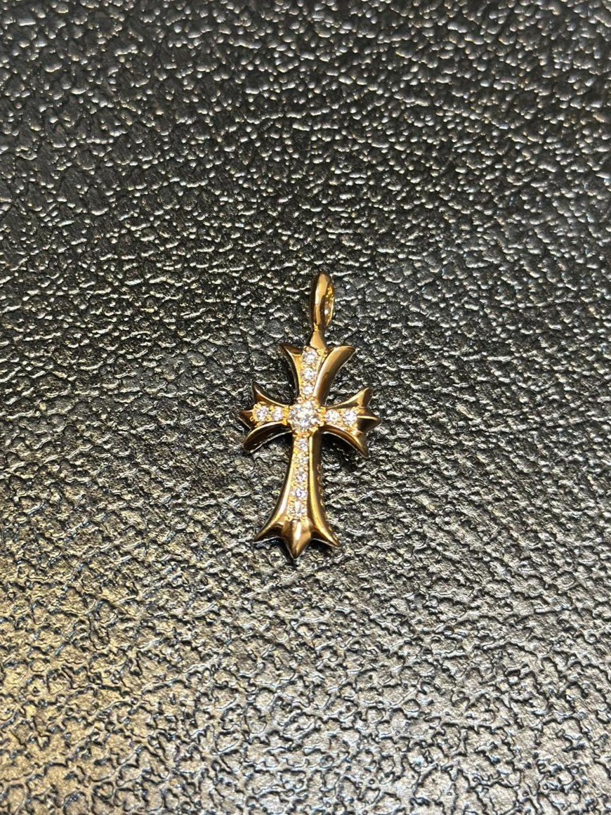 A gold cross pendant with small clear stones set along the edges, displayed on a textured dark gray surface.