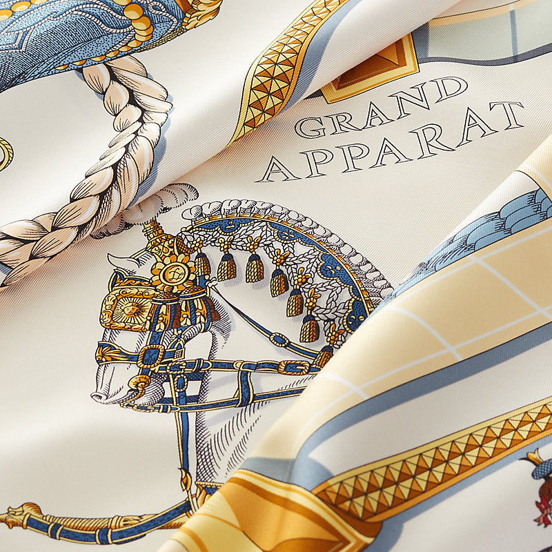 A close-up of a luxury silk scarf featuring a detailed print of a horse in ornate gold and blue harness, with the text 'GRAND APPARAT' printed on a cream background.