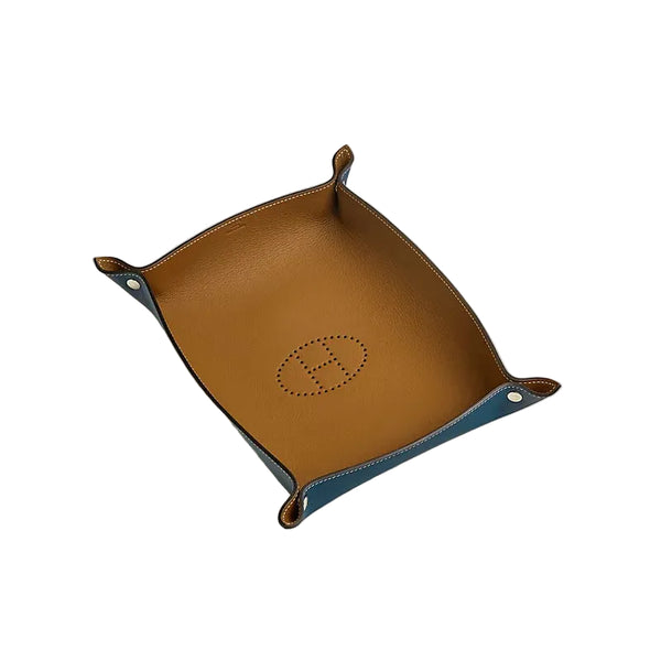 A square leather tray with a tan interior and blue exterior corners, featuring a debossed Hermès H logo in the center and white contrast stitching, photographed on a light gray background.
