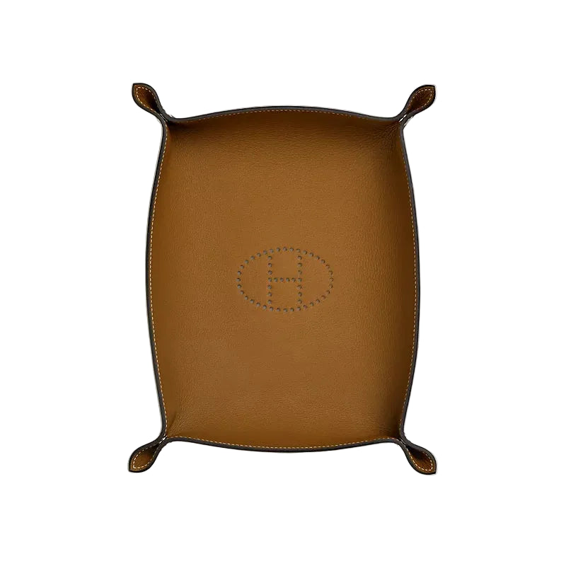 A brown leather Hermès tray with four small loop handles and a debossed oval H logo in the center, photographed from directly above on a light off-white background.