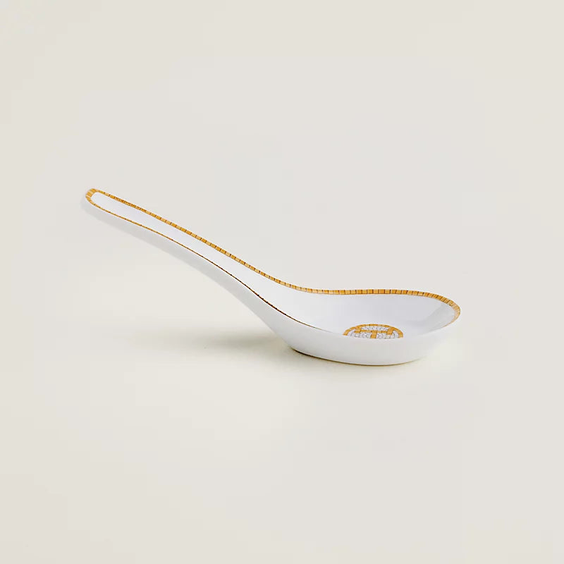 A white porcelain serving spoon with a delicate gold patterned rim and handle, featuring a small gold crest logo on the bowl, set against a solid off-white background.