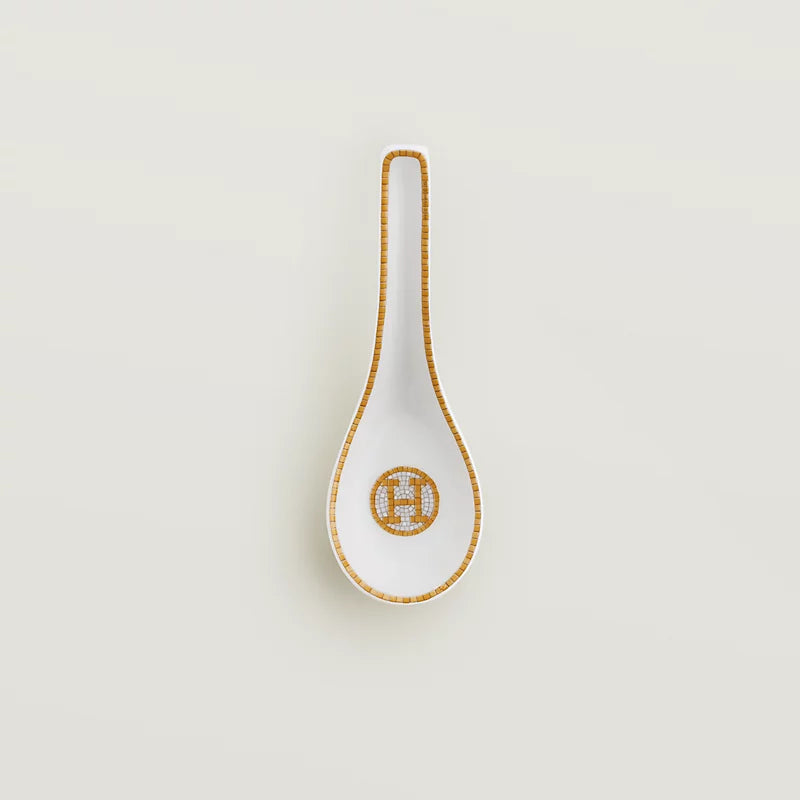 A white porcelain serving spoon with a gold geometric border and a gold monogram logo on the bowl, set against a solid light gray background.