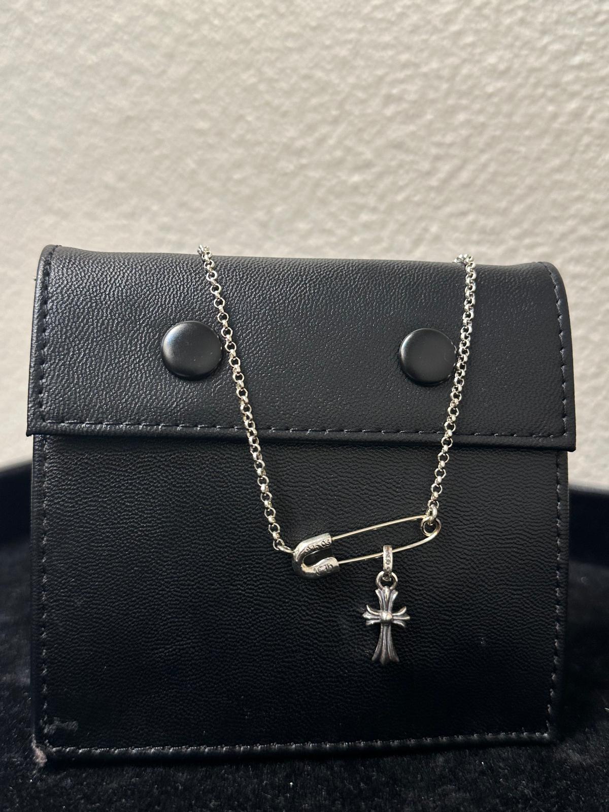 A silver safety-pin style pendant necklace with a small cross charm, displayed on a black faux-leather jewelry pouch against a textured off-white wall.